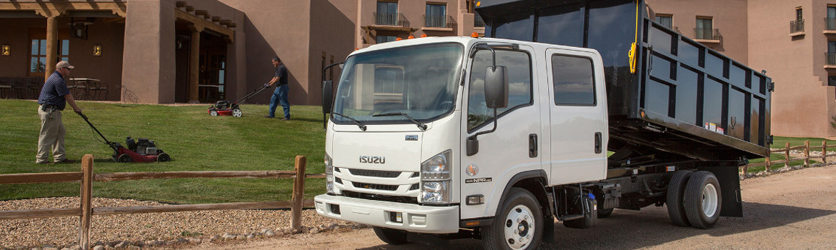 A white and black 2017 Isuzu Truck NPR Diesel sitting on a road next to workers mowing a lawn.